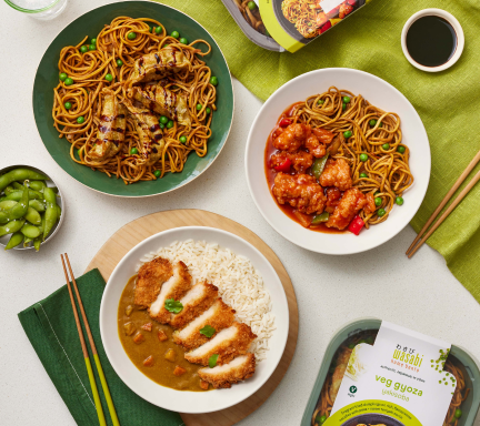 Top view of a table with three Asian-inspired dishes: noodles with grilled meat, spicy chicken with noodles, and curry with rice and sliced katsu. Edamame on the side. Packaging for veggie gyoza is visible, with green cloth napkins and chopsticks.