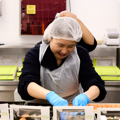 A woman wearing a hairnet and gloves smiles while preparing food in a kitchen. She is assembling sushi boxes. Another person is partially visible in the background, also wearing a hairnet. Sushi and packaging are on the counter.