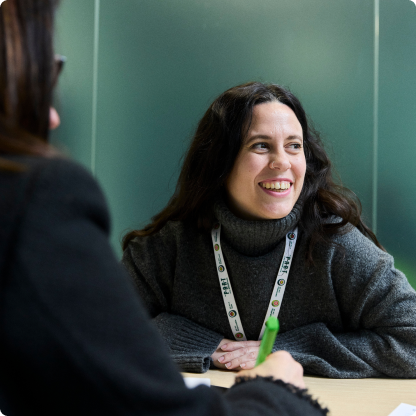 A woman with long dark hair, wearing a grey sweater and lanyard, smiles while sitting at a table. Another person is visible from the back in the foreground. The setting appears to be a casual meeting or discussion.