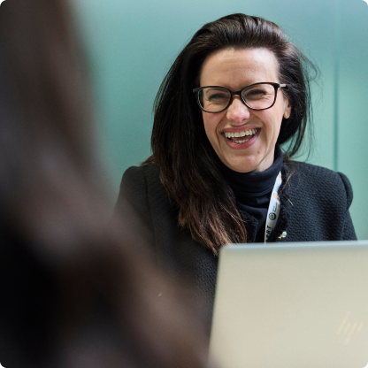 A person with long dark hair and glasses is smiling while sitting in front of a laptop. They are wearing a dark jacket and are in an indoor setting with a greenish background.
