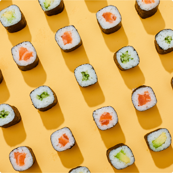 An overhead view of sushi rolls neatly arranged in a diagonal pattern on a bright yellow background. The rolls are filled with various ingredients like salmon, avocado, and cucumber, wrapped in seaweed.
