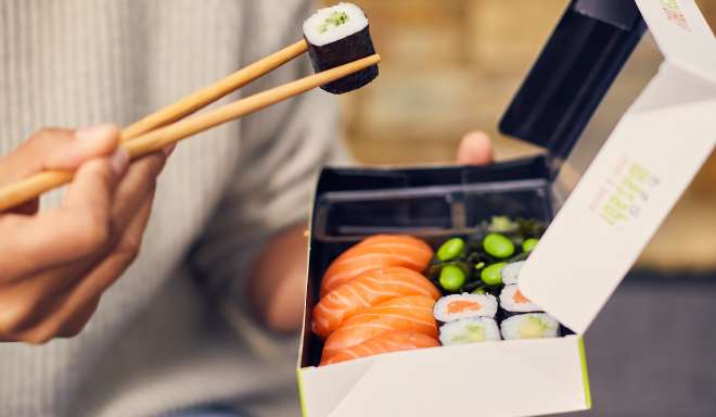 A person holding a sushi roll with chopsticks above a takeout box containing salmon nigiri, sushi rolls, and edamame. The person is wearing a gray sweater.