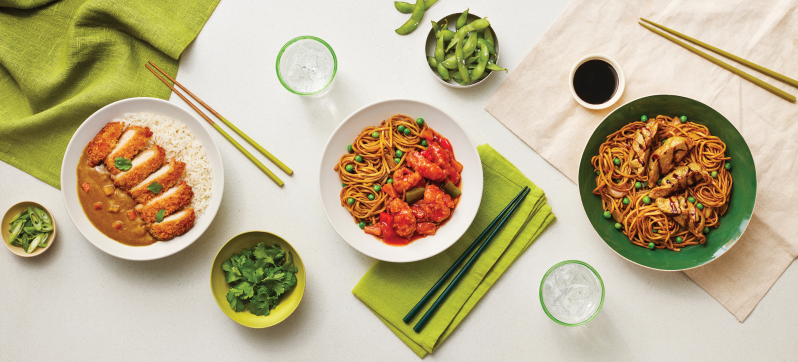 An overhead view of three dishes: a bowl with rice and curry sauce topped with sliced chicken, another bowl with noodles and vegetables in a red sauce, and a green bowl with noodles and grilled meat. Edamame, cilantro, and drinks are on the side.