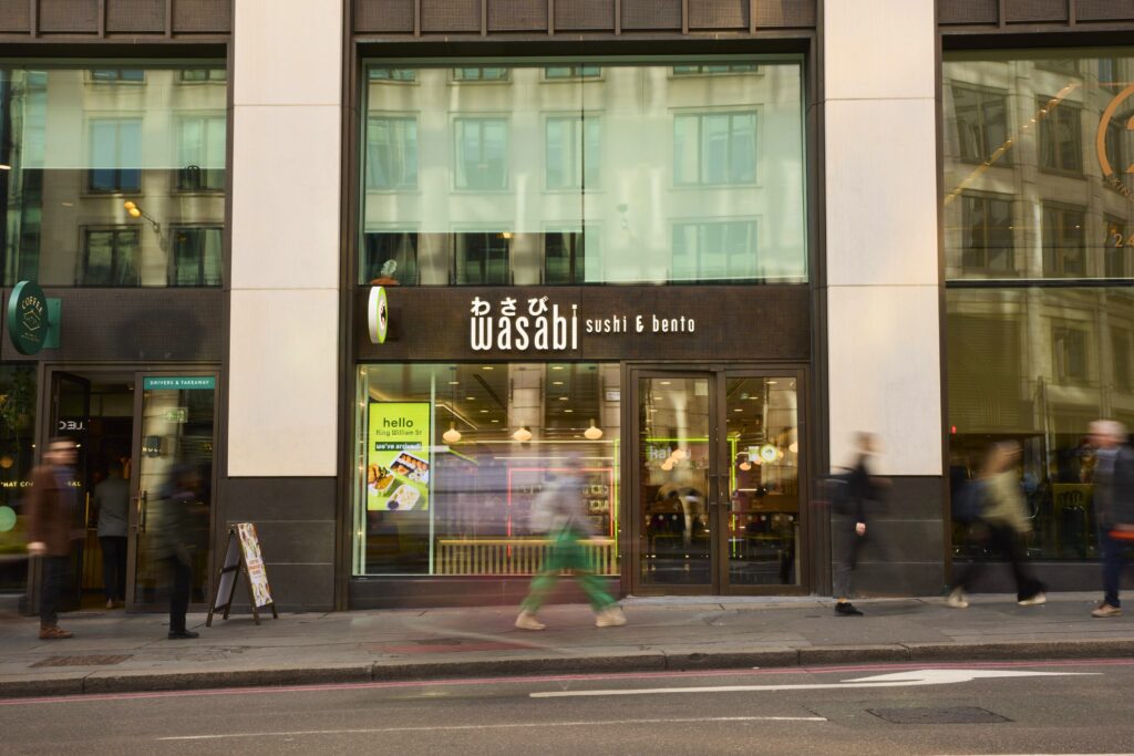 A street view of the Wasabi Sushi & Bento restaurant with large glass windows. Several blurred pedestrians walk past, and the building's reflection is visible in the windows.