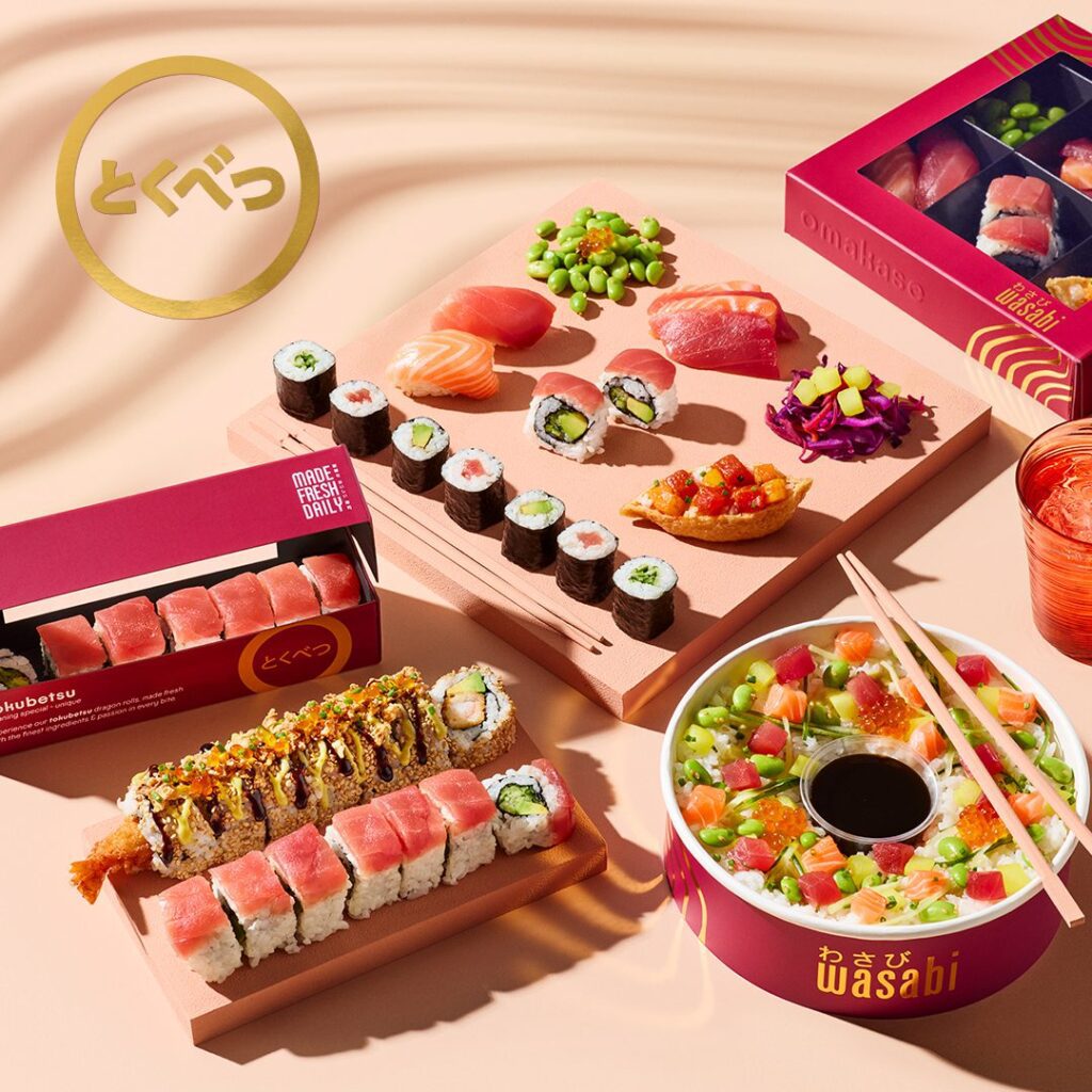A selection of sushi rolls, nigiri, sashimi, edamame, and poke in vibrant trays and bowls on a pastel pink surface, with branded packaging and chopsticks arranged neatly.