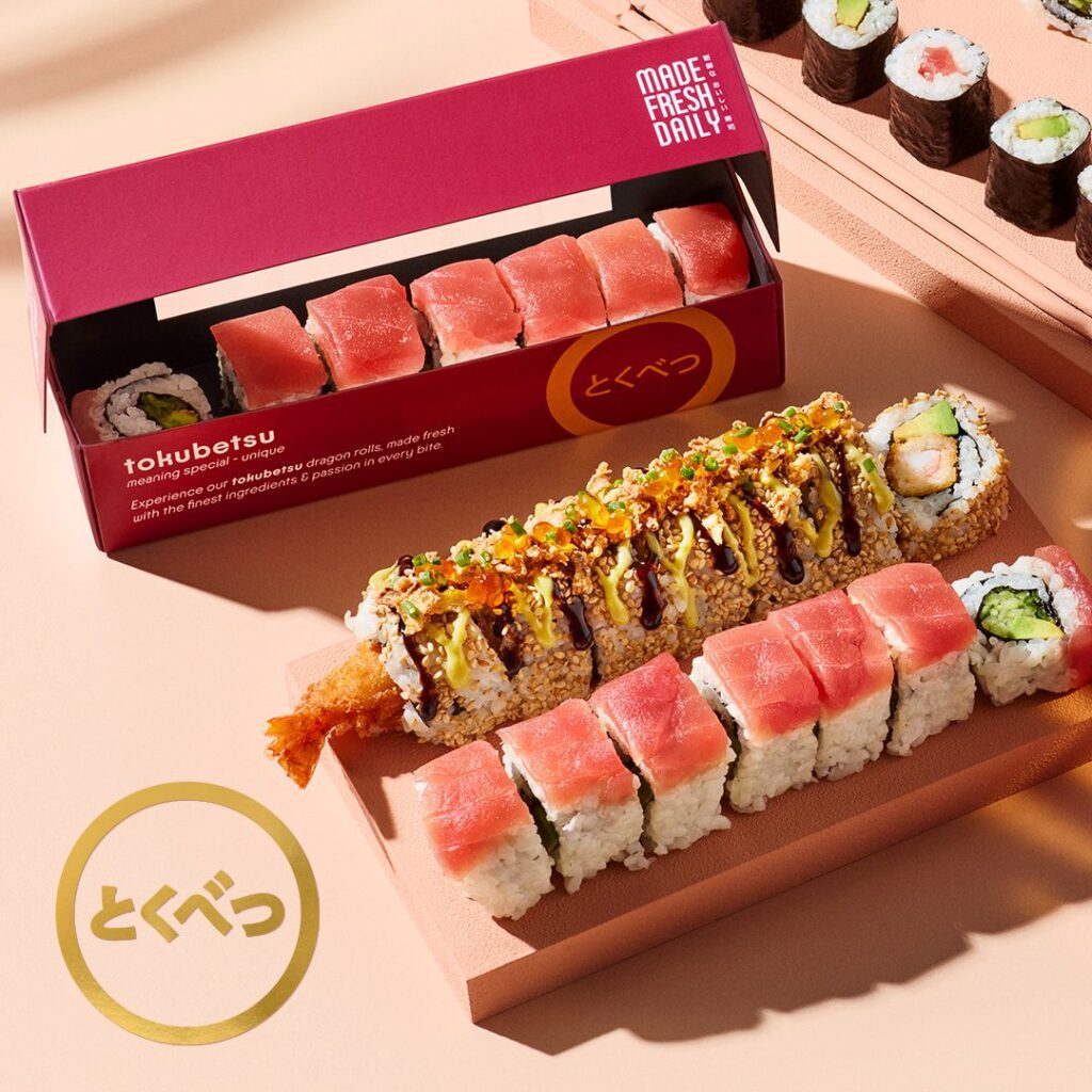 A vibrant display of assorted sushi rolls on wooden trays, including tuna-topped rolls, a tempura shrimp roll with sauce and garnish, and a boxed set labeled “tokubetsu,” all arranged on a light pink surface.
