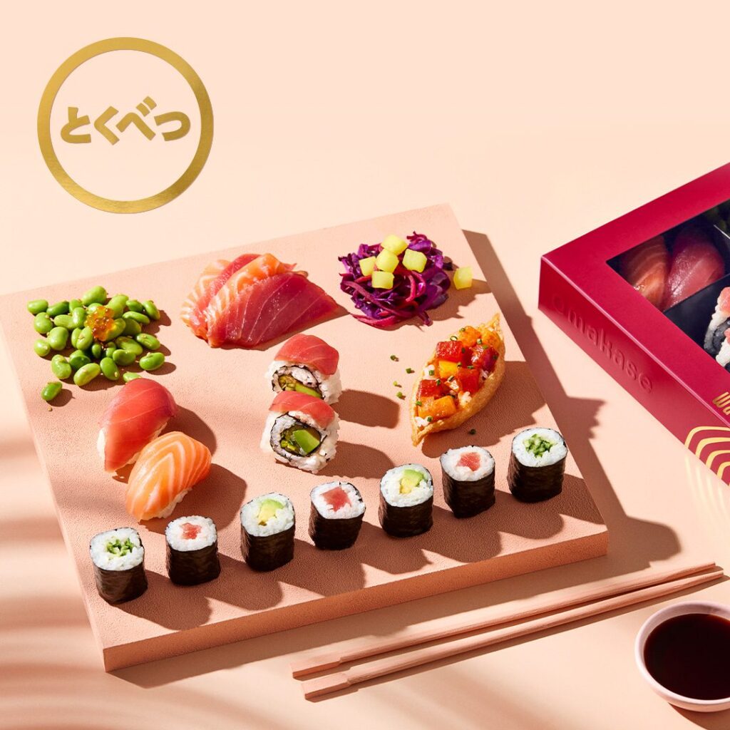 A sushi platter with assorted nigiri, sashimi, maki rolls, edamame, cabbage slaw, and poke on a square plate, next to chopsticks, soy sauce, and a pink sushi box on a peach-colored surface.