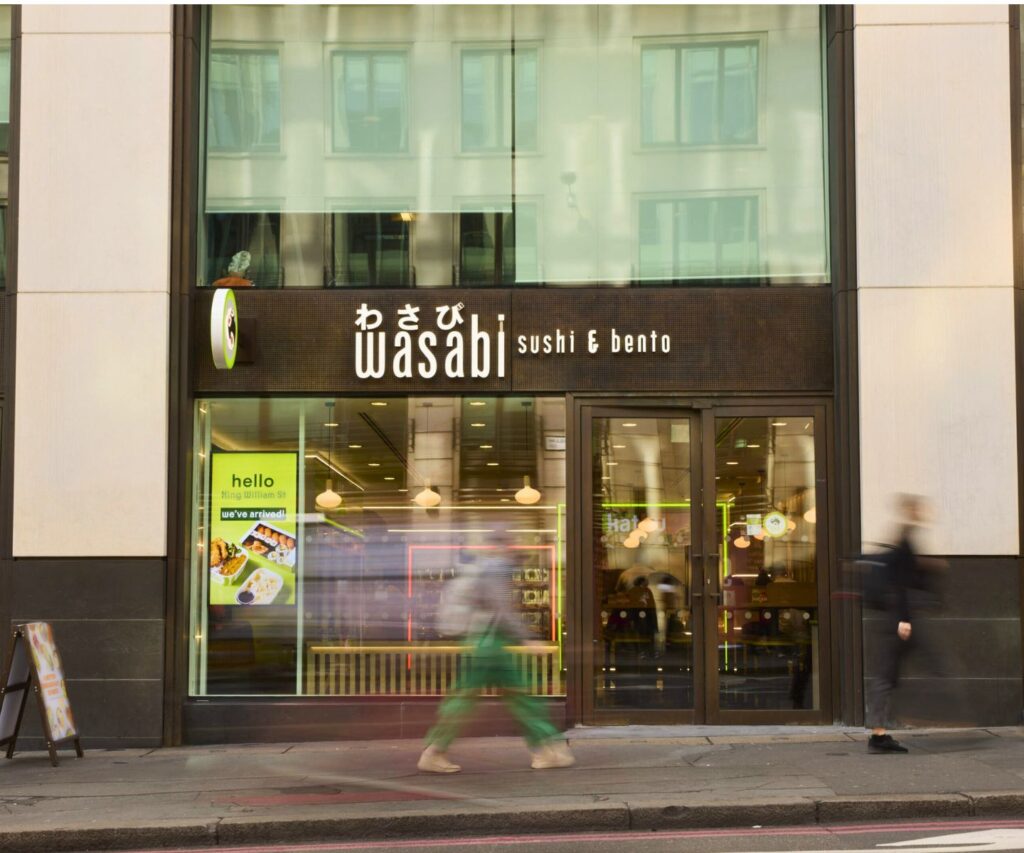 Storefront of Wasabi Sushi & Bento with illuminated signs, glass windows, and blurred pedestrians walking past on the sidewalk in an urban setting.