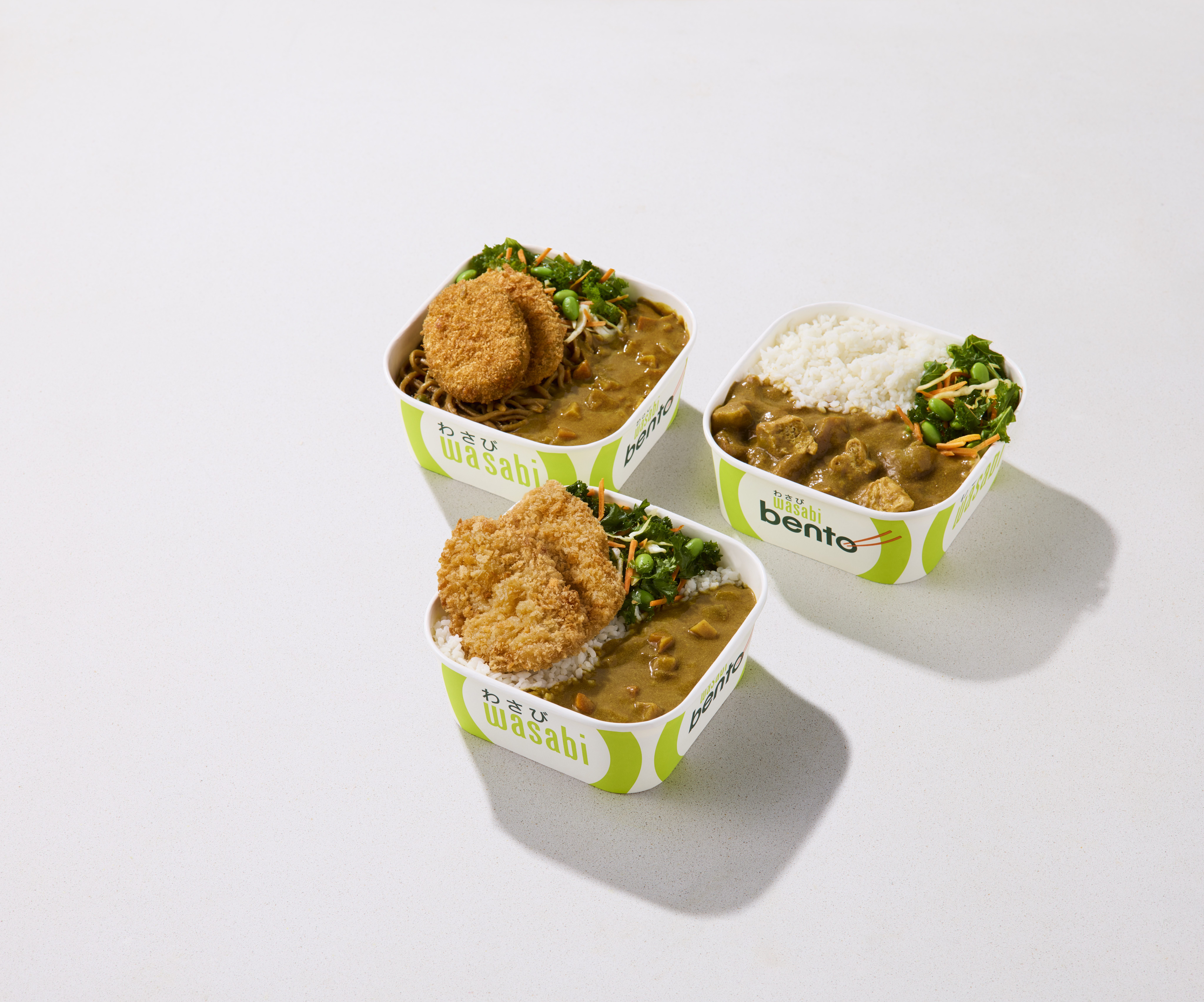 Three takeout bento boxes with white rice, curry, fried cutlets, and greens are arranged on a white surface. The boxes are labeled “wasabi” and “bento” with a green and white design.