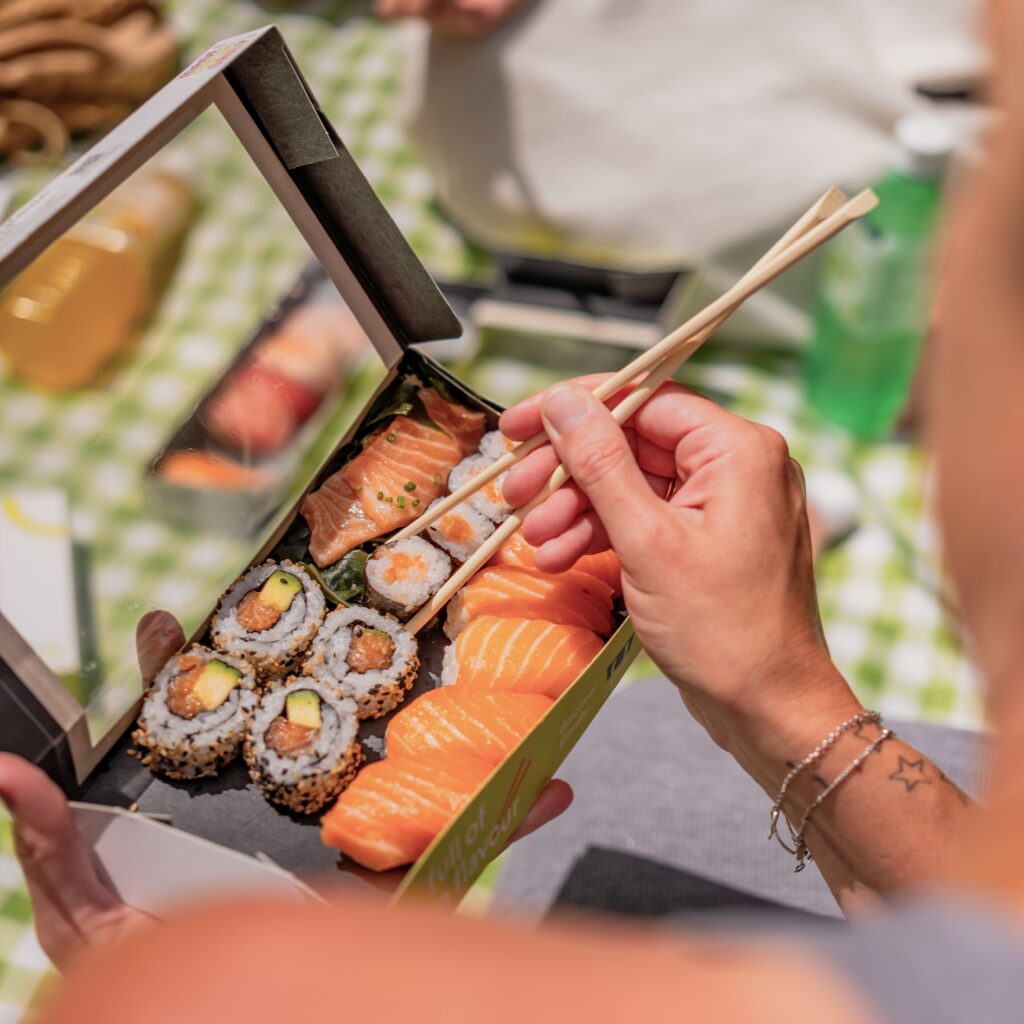 A person holding chopsticks is about to pick up a piece of sushi from a black box containing assorted sushi rolls and salmon sashimi, set on a checkered tablecloth.