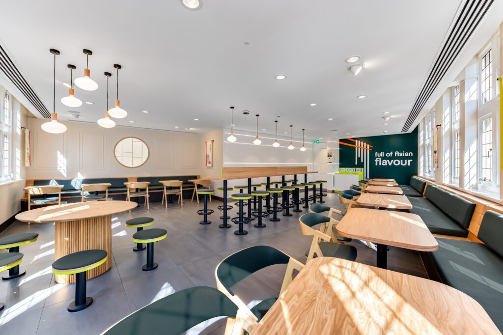A modern, brightly lit restaurant interior with wooden tables, green chairs and stools, and wall art reading 
