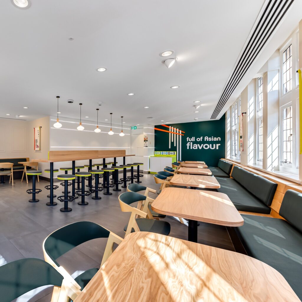 Bright, modern restaurant interior with wooden tables, green chairs, barstools at a counter, large windows, and a wall sign reading 