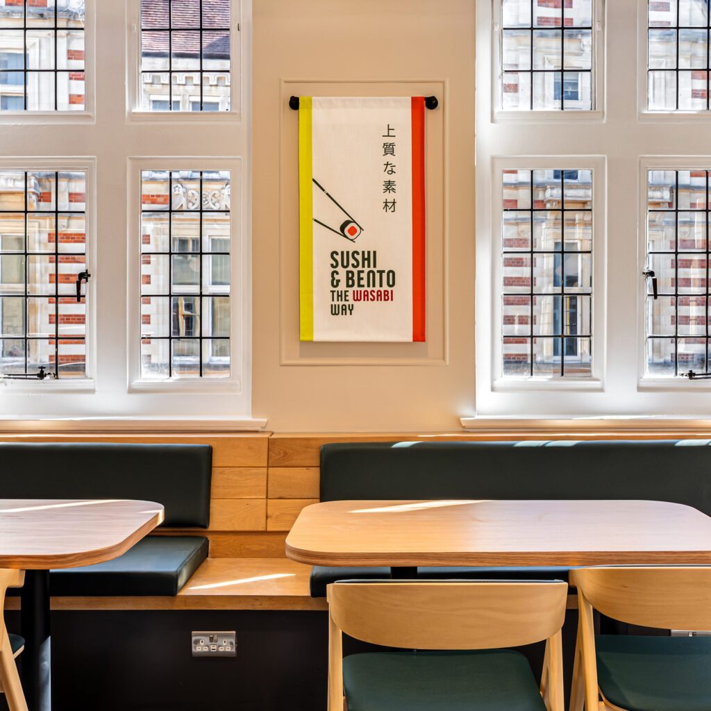 A bright, modern dining area features wooden tables, green cushioned seats, tall windows, and a decorative banner reading “Sushi & Bento The Wasabi Way” hanging on the wall.
