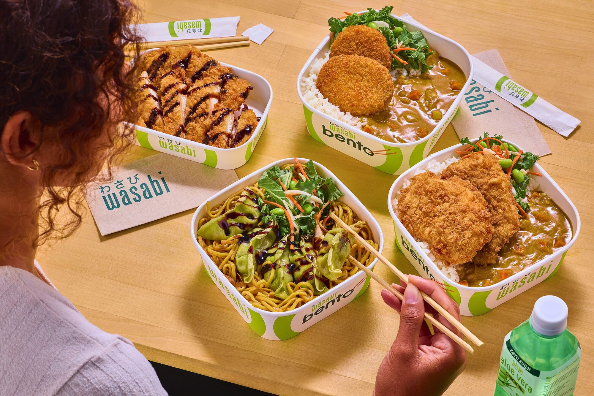 A person holds chopsticks over a bento box with noodles and vegetables. Three other bento boxes with curry, rice, katsu, and croquettes are on the table, along with Wasabi-branded napkins and a bottle of water.