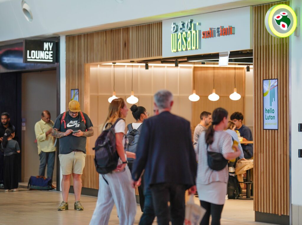 People walk past and queue at Wasabi Sushi & Bento in an airport terminal. The restaurant has a modern wooden exterior, with people eating inside. Signs for 