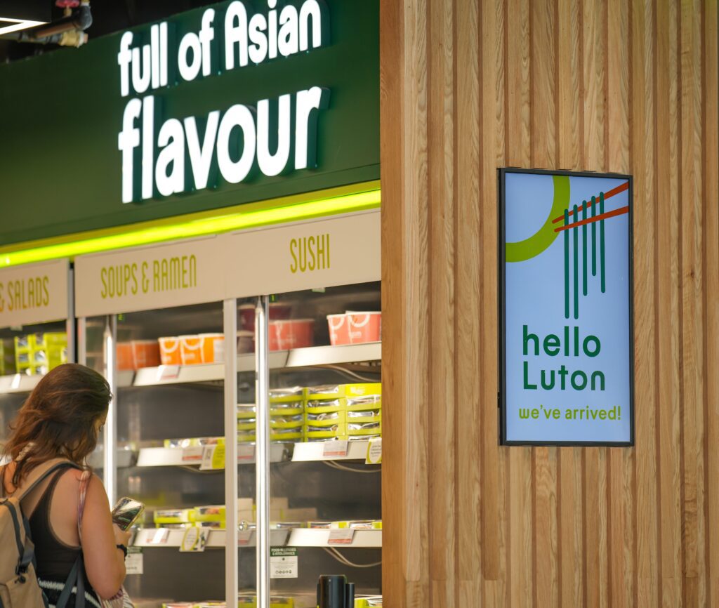 A woman stands near refrigerated shelves of Asian meals under a sign reading
