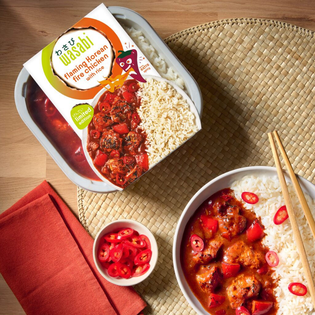 A bowl of rice and spicy red chicken curry is seen next to its packaging, which reads “wasabi flaming Korean fire chicken.” Sliced red chilies are in a small bowl, with chopsticks and a napkin on a woven placemat.