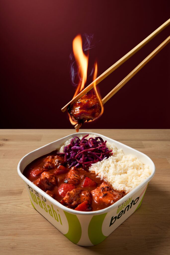 A pair of chopsticks holds a flaming piece of saucy chicken above a takeout container filled with rice, red cabbage, and more chicken, set on a wooden table against a dark red background.