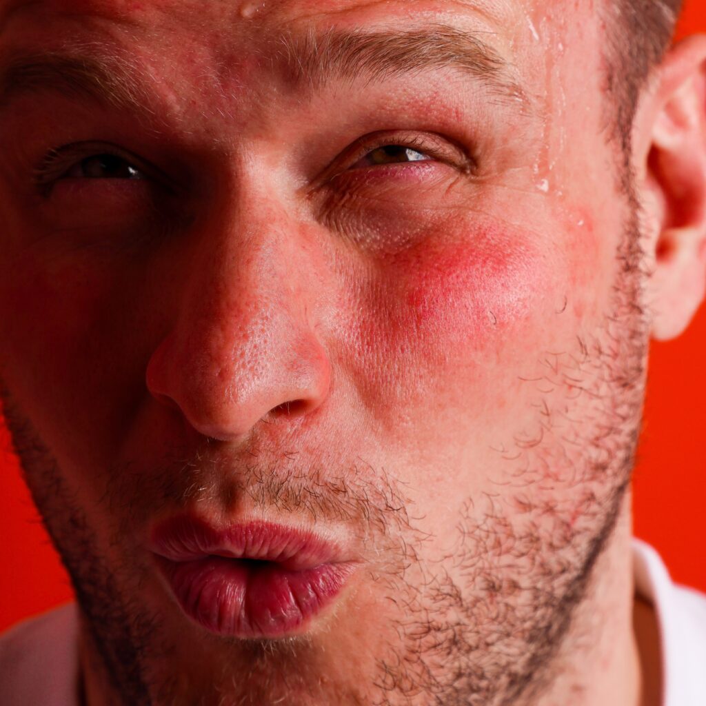A close-up of a man's face with flushed cheeks, pursed lips, squinting eyes, and beads of sweat on his forehead, set against a bright orange background. He appears to be reacting to something intense.