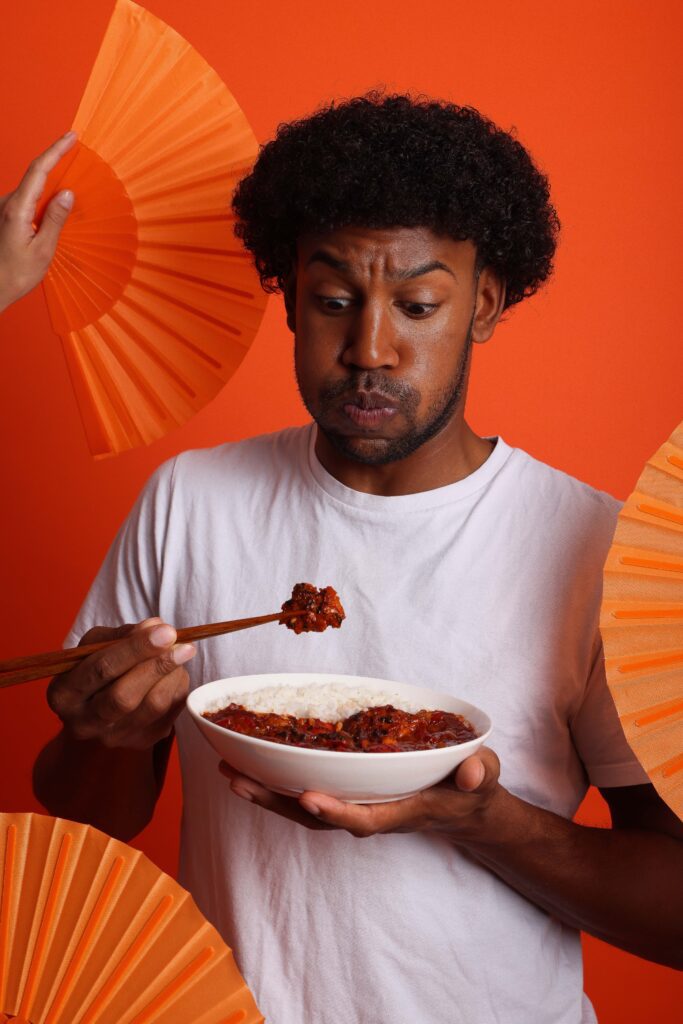 A man in a white shirt, holding a bowl of rice and spicy food with chopsticks, looks surprised. Orange fans frame him against a bright orange background.