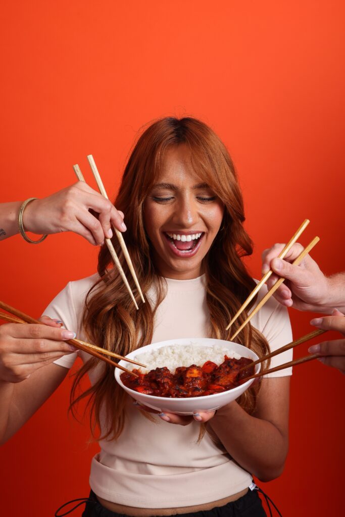 A smiling woman holds a bowl of rice and sauce while several hands with chopsticks reach toward the food against an orange background.