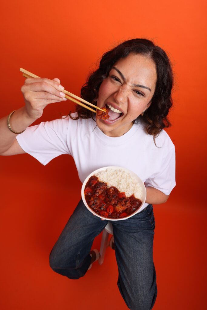 A woman with curly dark hair, wearing a white shirt and jeans, sits on a stool against an orange background. She is holding a bowl of rice with sauce and eating with chopsticks, smiling energetically at the camera.