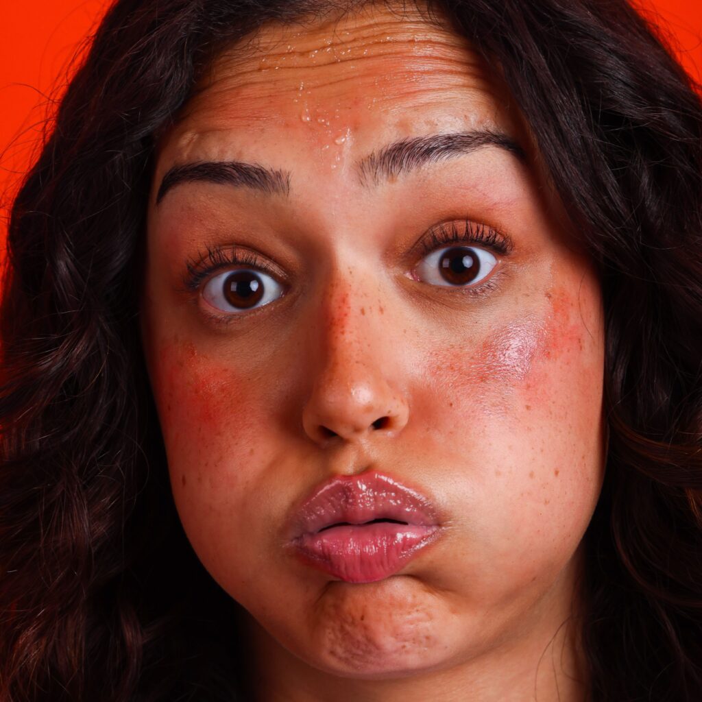 A woman with flushed cheeks and lips pursed, puffing her cheeks out. She has beads of sweat on her forehead and a wide-eyed, surprised expression. The background is bright orange.