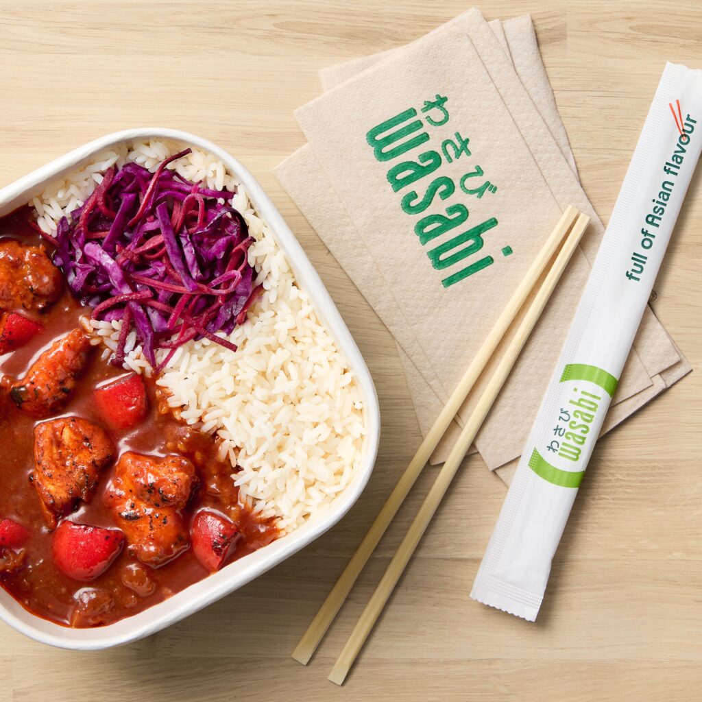 A bowl of rice with red sauced chicken and shredded purple cabbage, beside wooden chopsticks, napkins labeled 