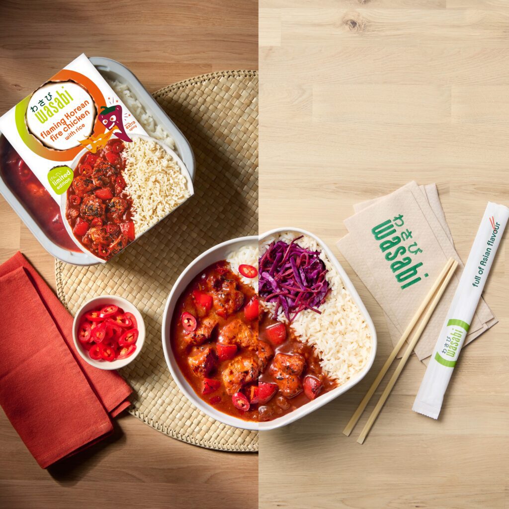 A bowl of mango chicken curry with rice and red cabbage sits on a woven mat beside its packaging, chopsticks, a Wasabi-branded napkin, sliced red chili, and an orange napkin on a light wooden surface.