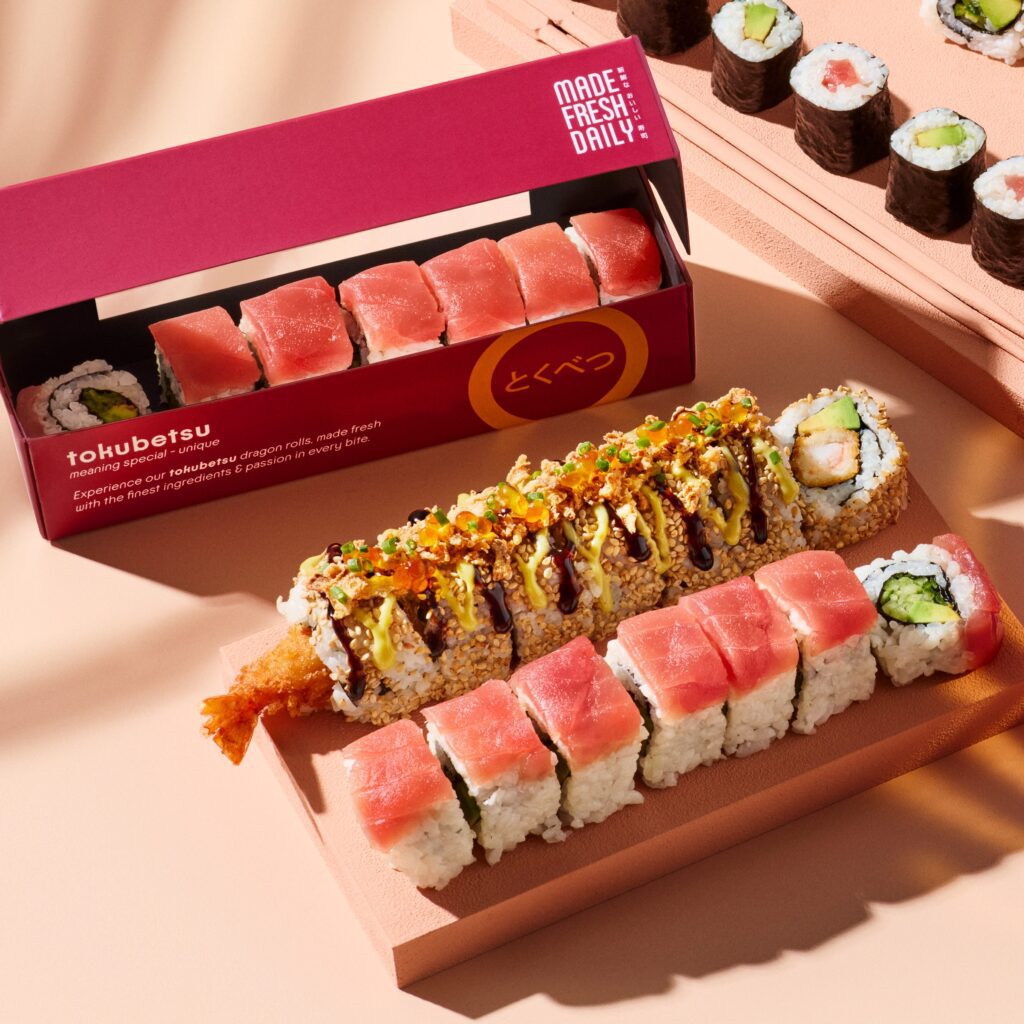 A selection of sushi rolls on light wooden trays, including tuna, cucumber, and tempura rolls. One roll is drizzled with sauces and toppings. A red box labeled “tokubetsu” holds more tuna sushi pieces.