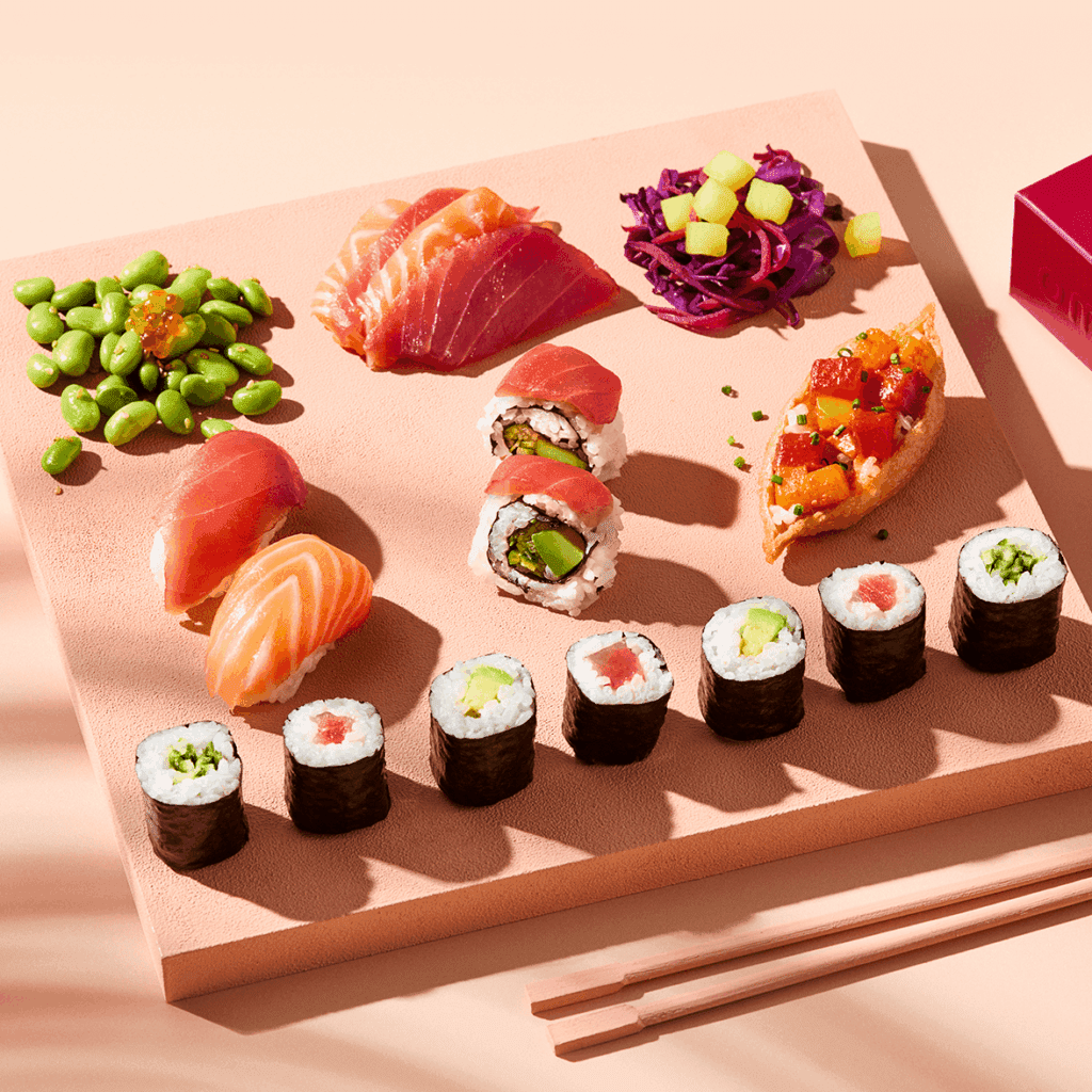 A variety of sushi, including nigiri, maki rolls, sashimi, and poke, is arranged neatly on a pink square plate with chopsticks beside it. Edamame and a red cabbage salad are also included.