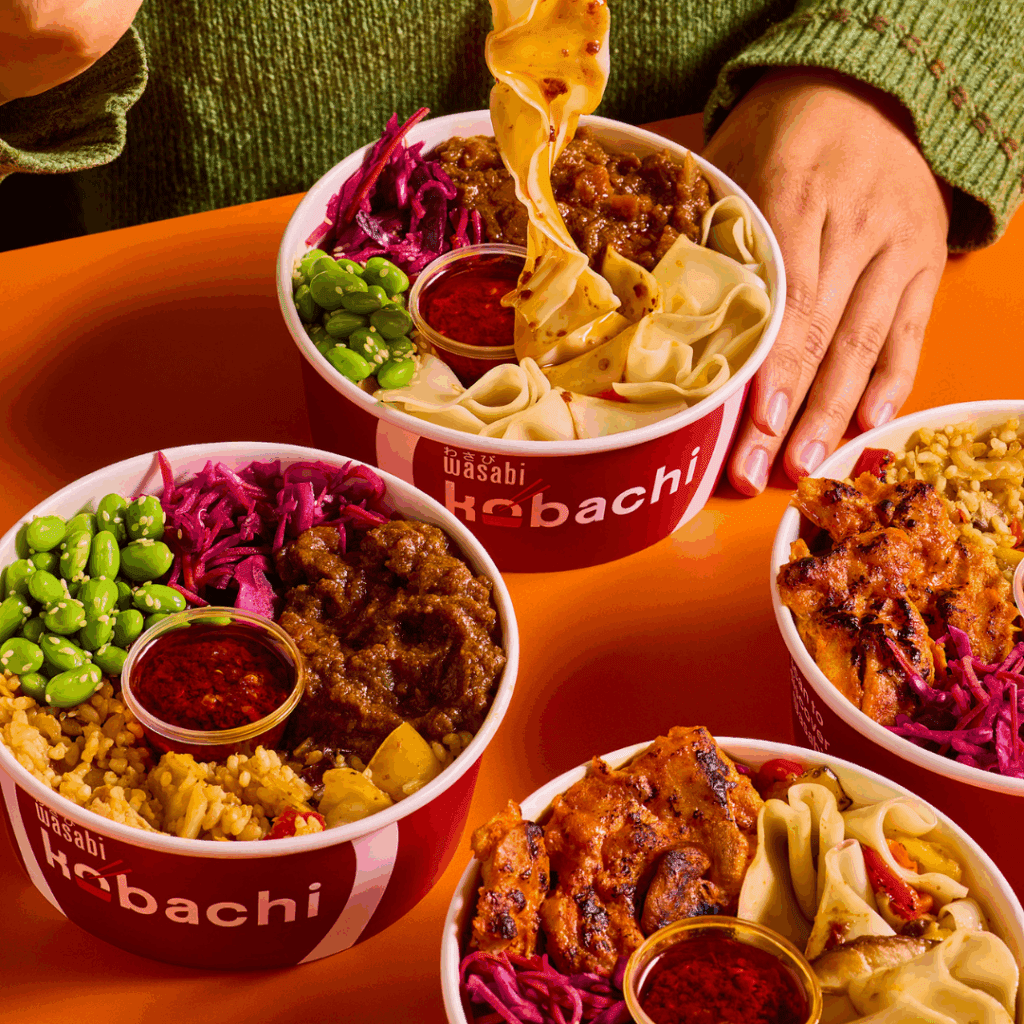 Three bowls of food with red Wasabi Hibachi branding, filled with rice, noodles, vegetables, meat, and sauce. A hand wearing a green sleeve is picking up noodles from one of the bowls. The background is orange.