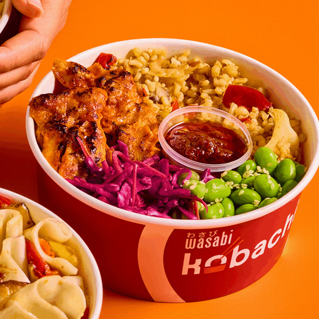 A vibrant rice bowl with grilled chicken, red cabbage, edamame, brown rice, vegetables, and a small container of red sauce, served in a red container labeled