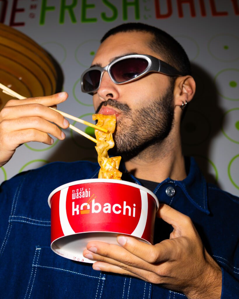 A man wearing sunglasses and a blue shirt eats noodles with chopsticks from a red and white Kobachi takeout container. He is mid-bite, holding the container close to his mouth.