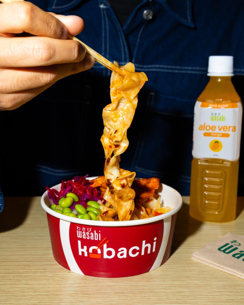 A hand lifts wide noodles with chopsticks from a red Kobachi bowl filled with colorful vegetables. A bottle of aloe vera drink stands in the background on a wooden table.