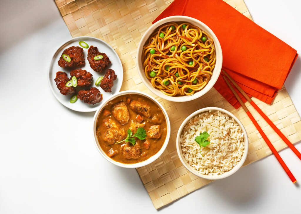 Three bowls of food containing noodles with green onions, white rice with cilantro, and chicken curry are arranged on a woven mat beside orange napkins and chopsticks, with a plate of glazed chicken bites.
