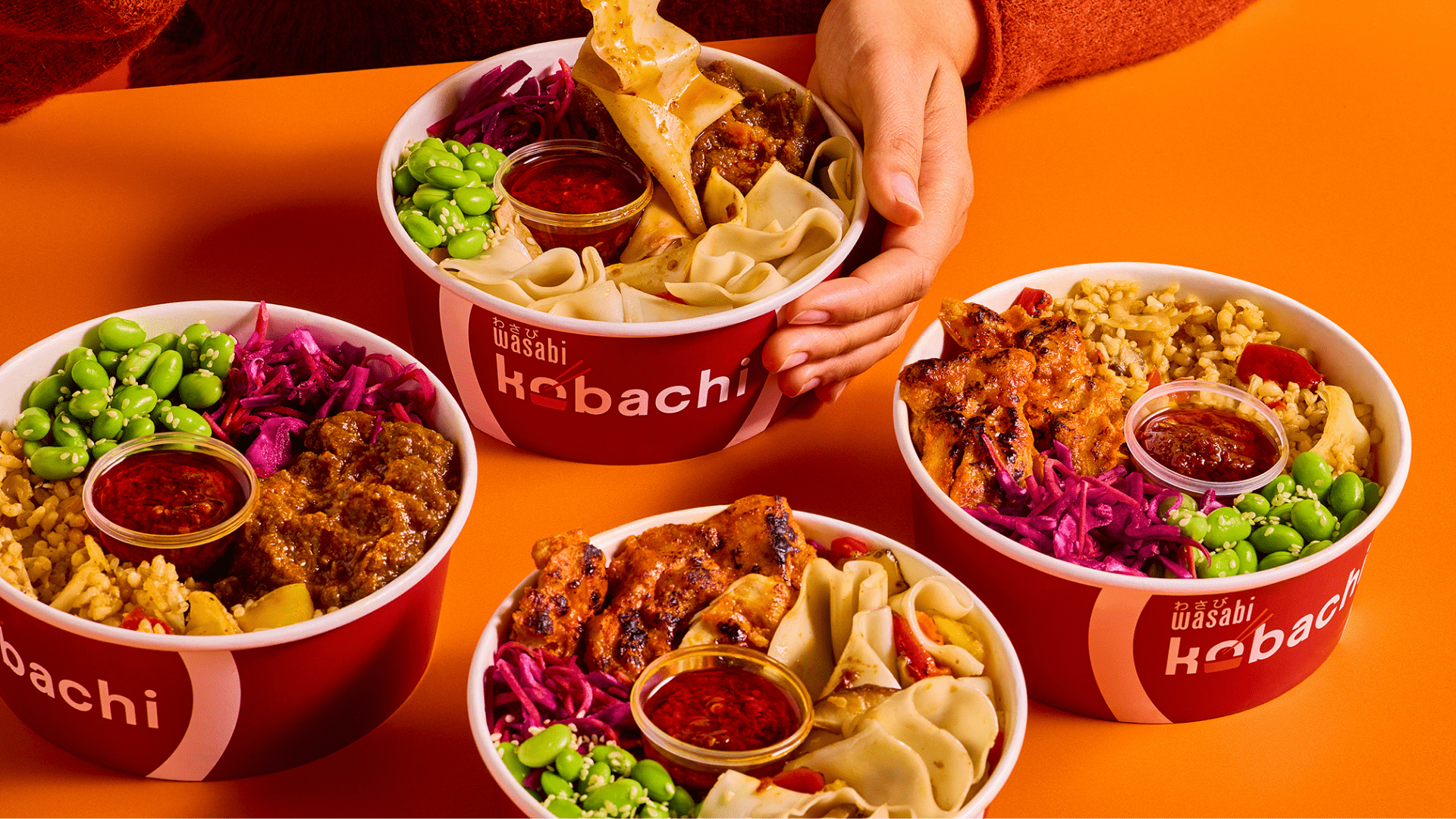 Four colorful bowls of Hibachi food with rice, grilled meat, edamame, purple cabbage, dumplings, and dipping sauce on an orange table. A hand is holding one of the bowls.