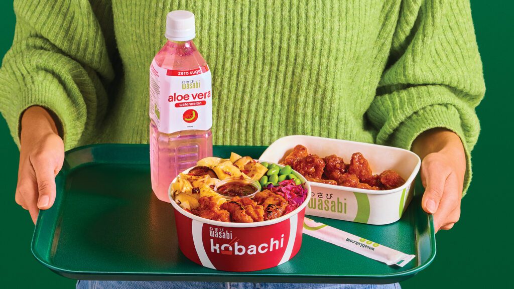 A person in a green sweater holds a dark green tray with a bottle of aloe vera drink, a bowl of rice with chicken, edamame, and vegetables, a side of fried chicken, and a wasabi packet.