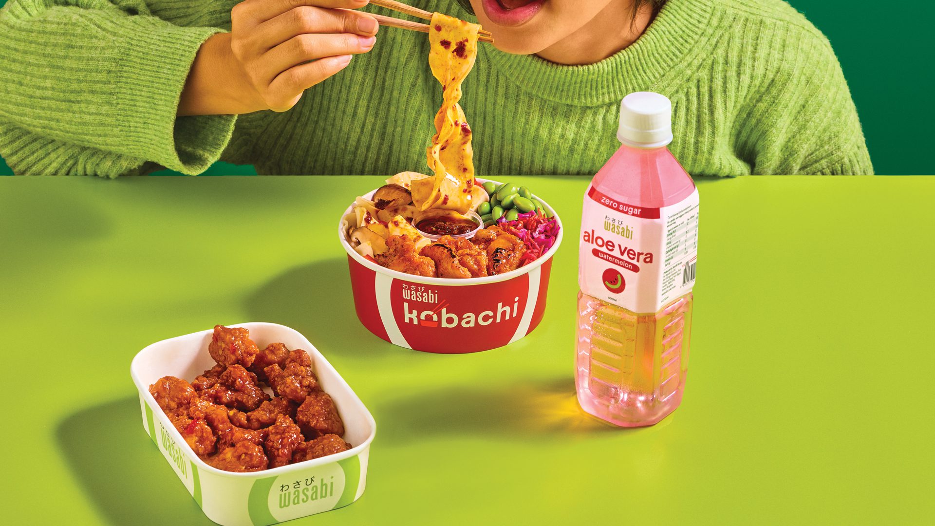 A person in a green sweater eats noodles with chopsticks from a bowl labeled “Kobachi.” Next to the bowl are a container of fried chicken and a pink bottle of aloe vera drink on a green table.