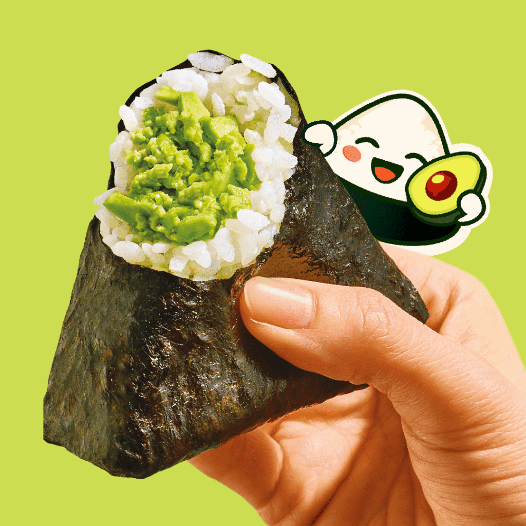 A hand holds a seaweed-wrapped rice ball (onigiri) with avocado filling. In the background, there's a cute cartoon onigiri, smiling and holding an avocado, on a light green backdrop.