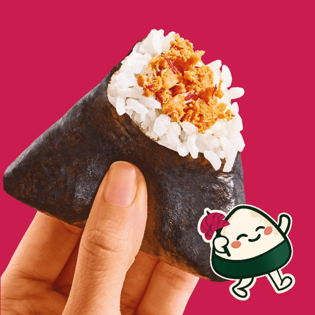 A hand holds a triangular rice ball wrapped in seaweed with a spicy filling on top. A cute cartoon onigiri character waves and holds a red heart in the bottom right corner. The background is solid pink.