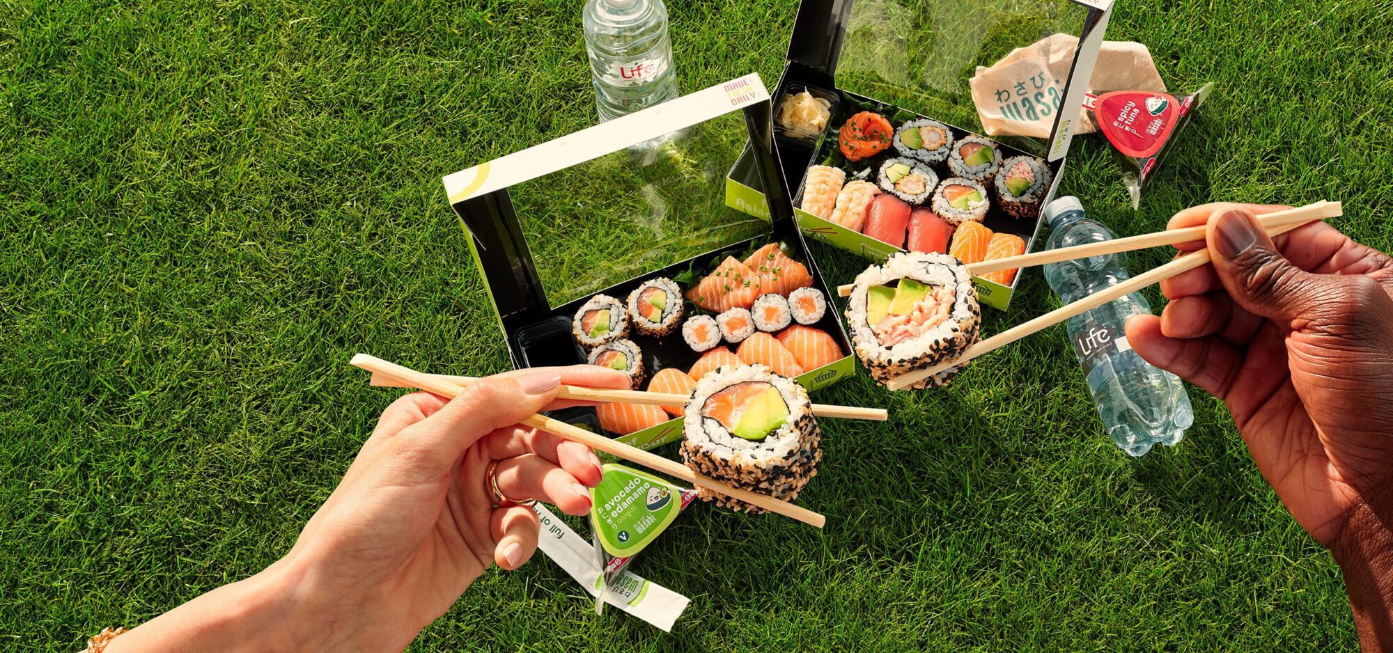 Two people use chopsticks to pick up sushi rolls and sample new onigiri flavours from takeaway sushi boxes on green grass, with bottled water, soy sauce, and serviettes nearby, suggesting a picnic setting.