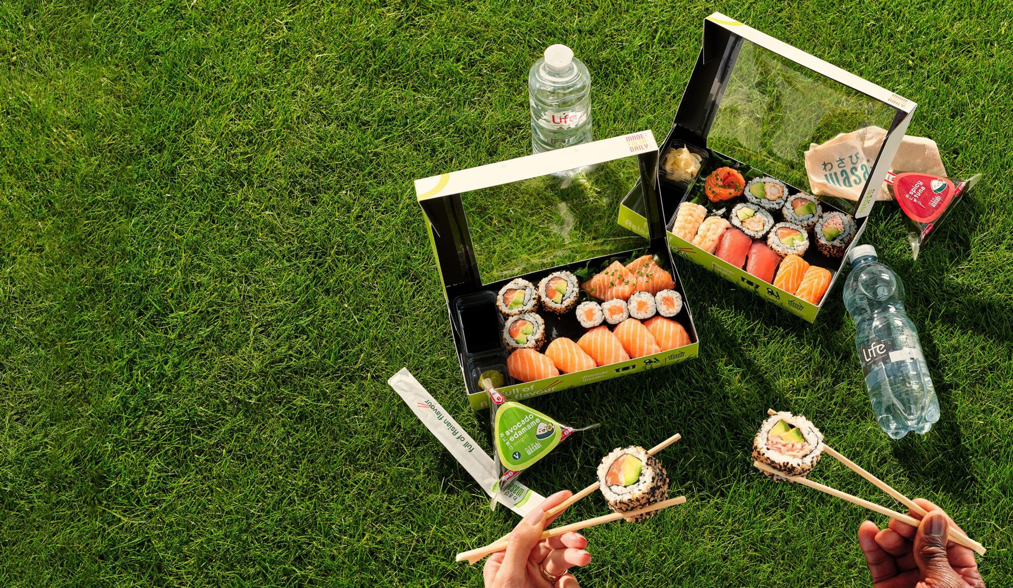 Two open sushi boxes, chopsticks, soy sauce, serviettes, and bottled water are arranged on green grass. Two hands hold sushi rolls with chopsticks in the foreground, suggesting a picnic setting.
