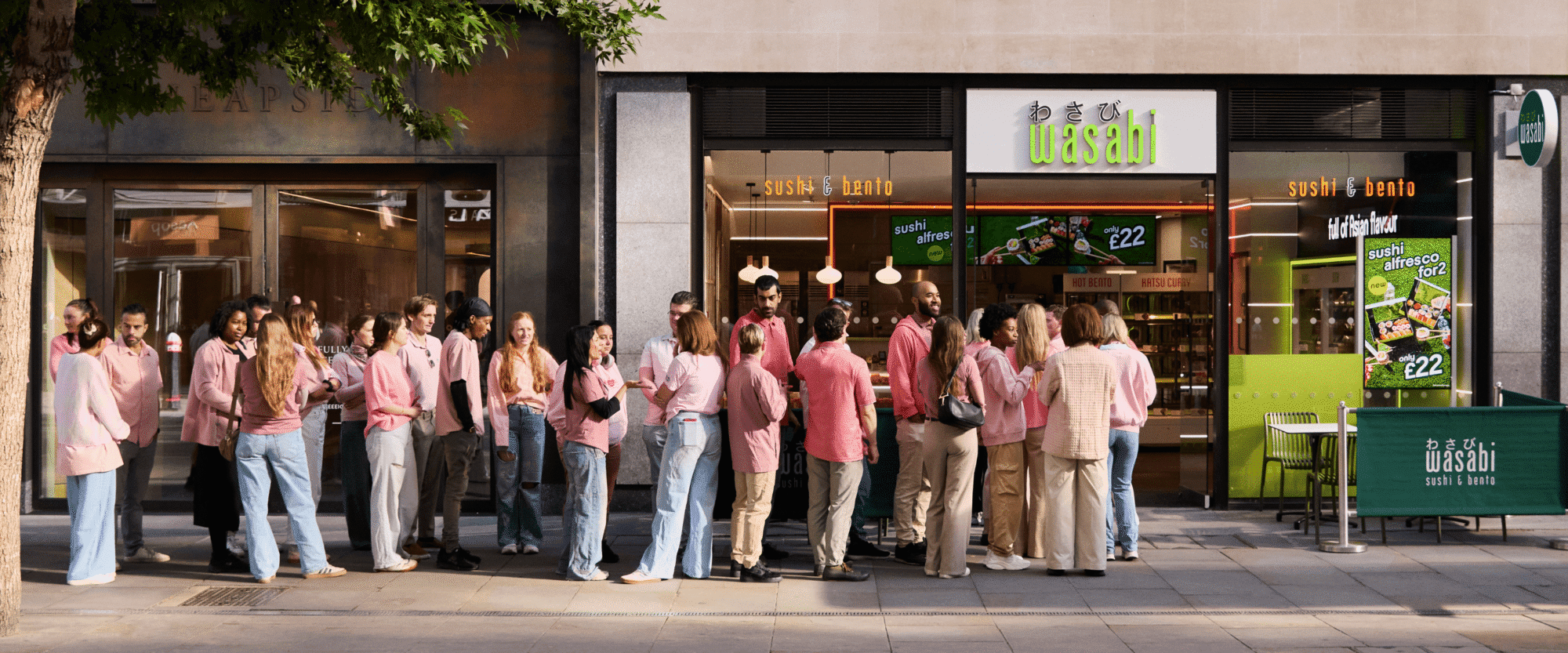 A group of people wearing pink tops and jeans queues up outside a Wasabi sushi and bento restaurant on a city street, with some people talking and others looking at the restaurant.