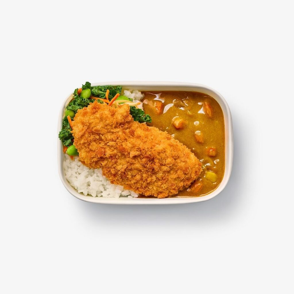 A rectangular dish with white rice, green vegetables, and a breaded fried cutlet topped with golden curry sauce containing peas and carrots.