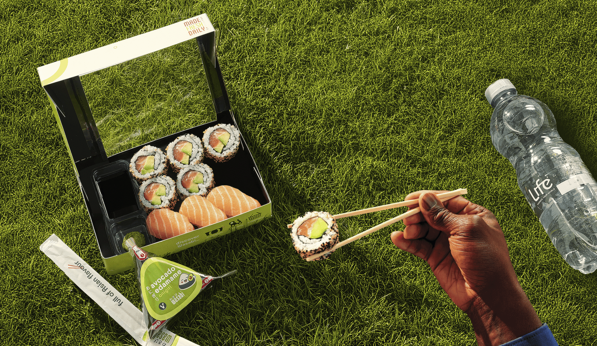 A hand holds sushi with chopsticks over a sushi box on grass, next to soy sauce, wasabi, ginger, and a bottle of water.