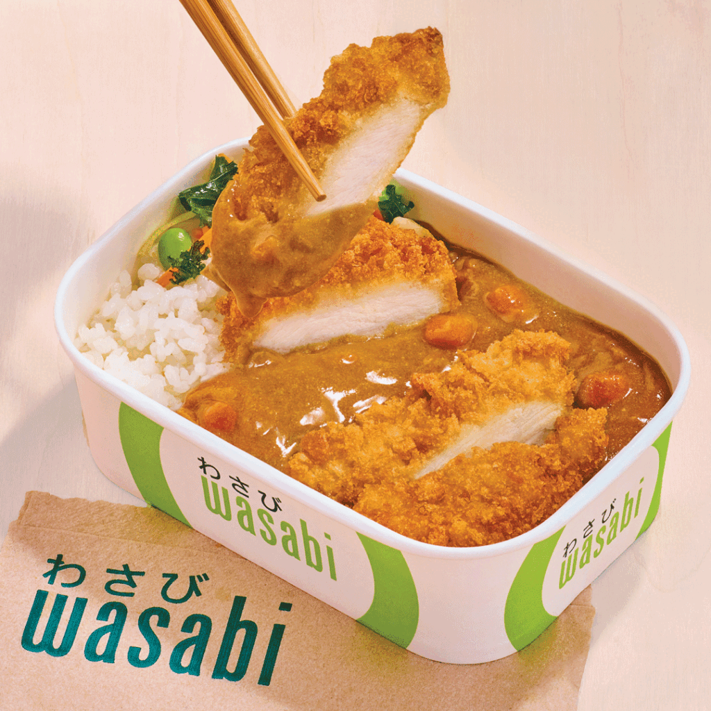 A takeaway container labelled “wasabi” holds white rice, curry with vegetables, and crispy breaded chicken. Chopsticks lift a sliced piece of chicken above the container. A serviette with “wasabi” is nearby.